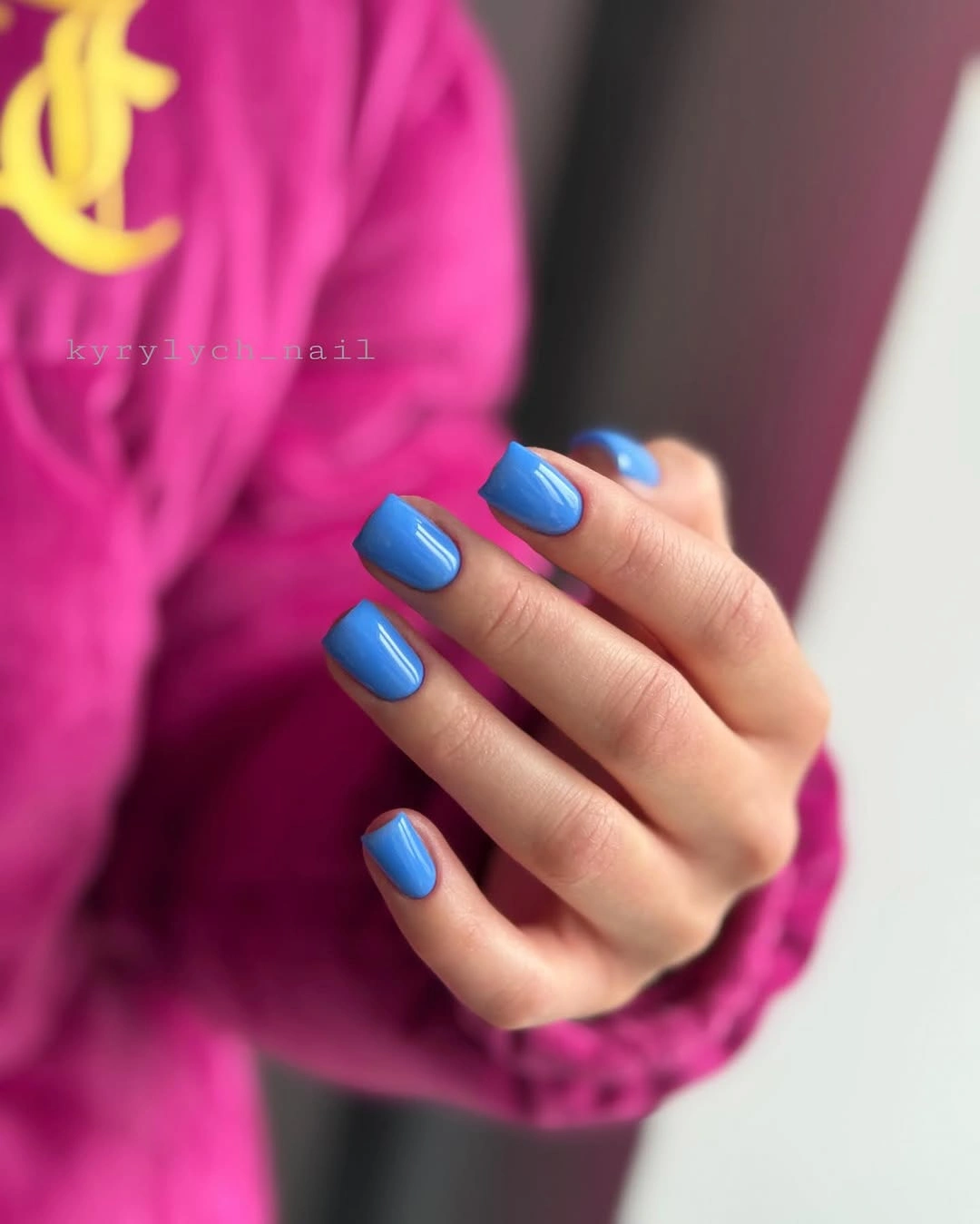 Glossy bright blue square nails against a vivid pink sweater
