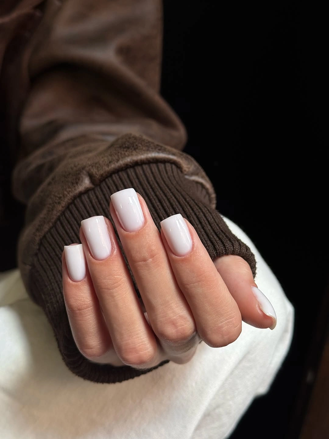 Short glossy white square nails against a dark knit sleeve