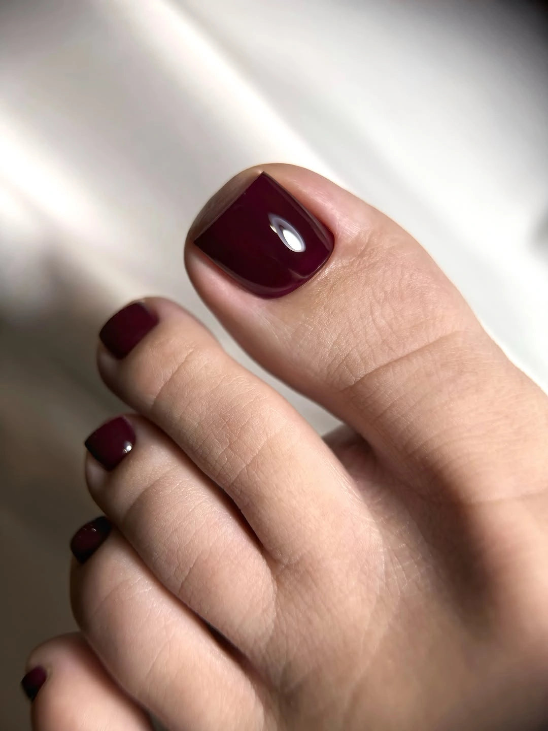 Short glossy burgundy square toenails against white fabric