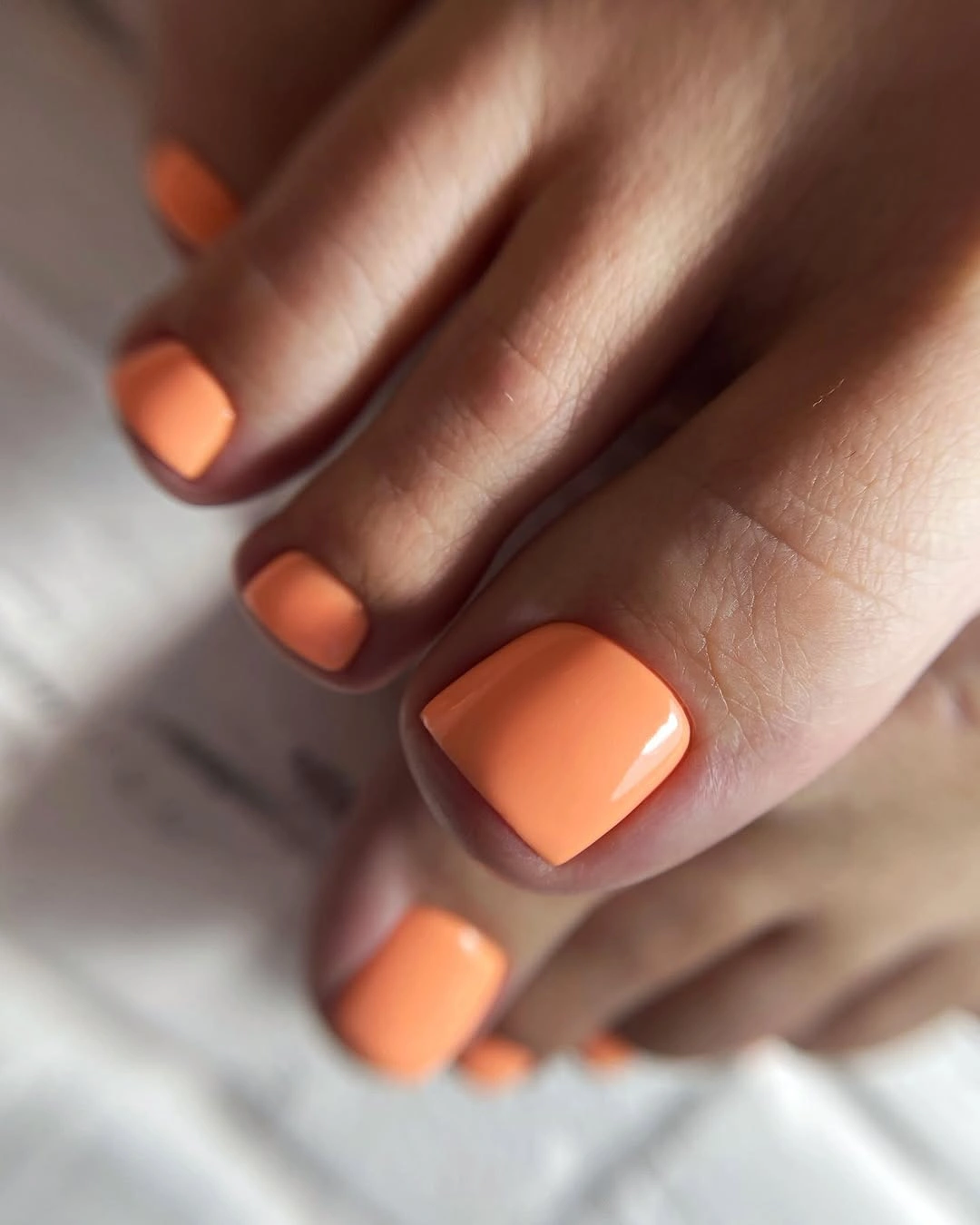 Short glossy peach-orange square toenails in soft light
