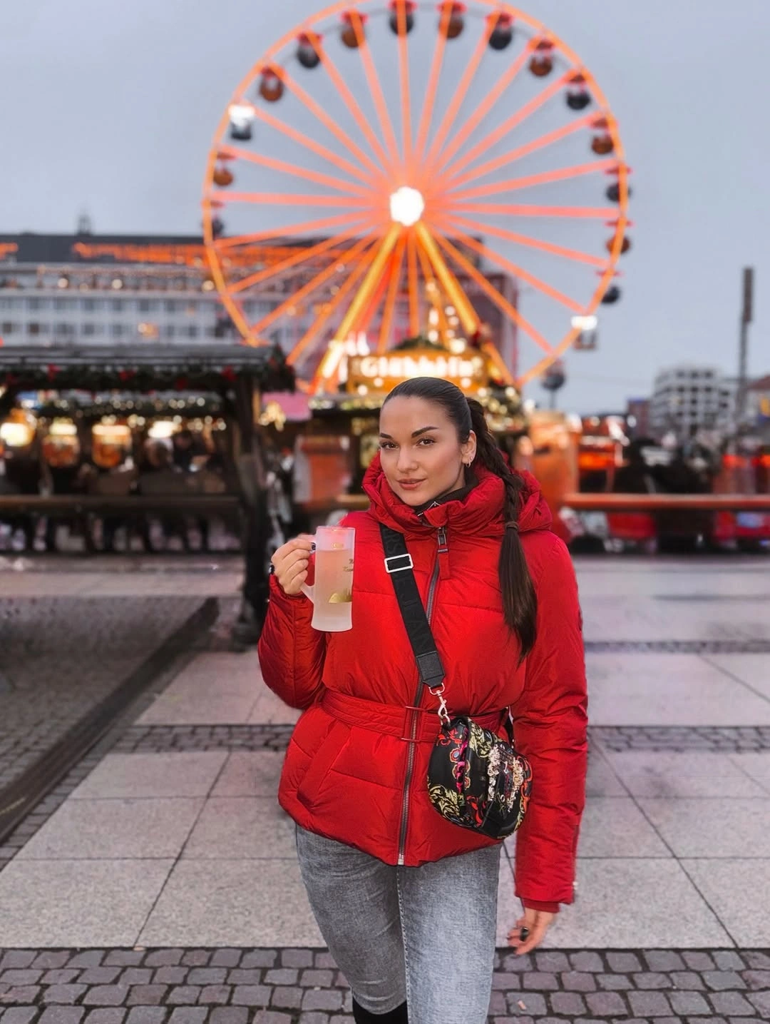 Red puffer-style jacket, gray pants, dark crossbody bag, and drink