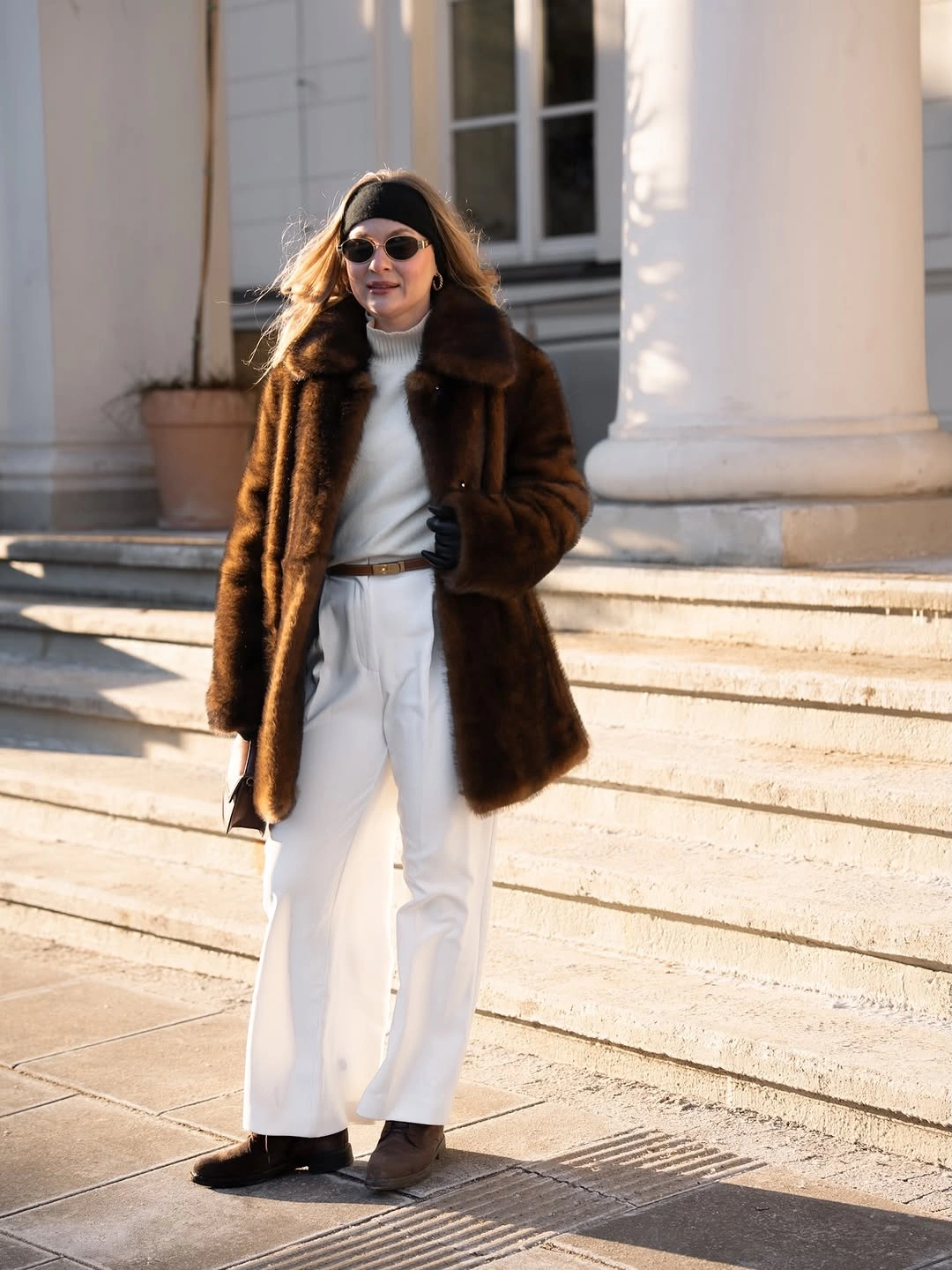 Brown plush-looking coat, white top and trousers, belt, sunglasses, and dark shoes