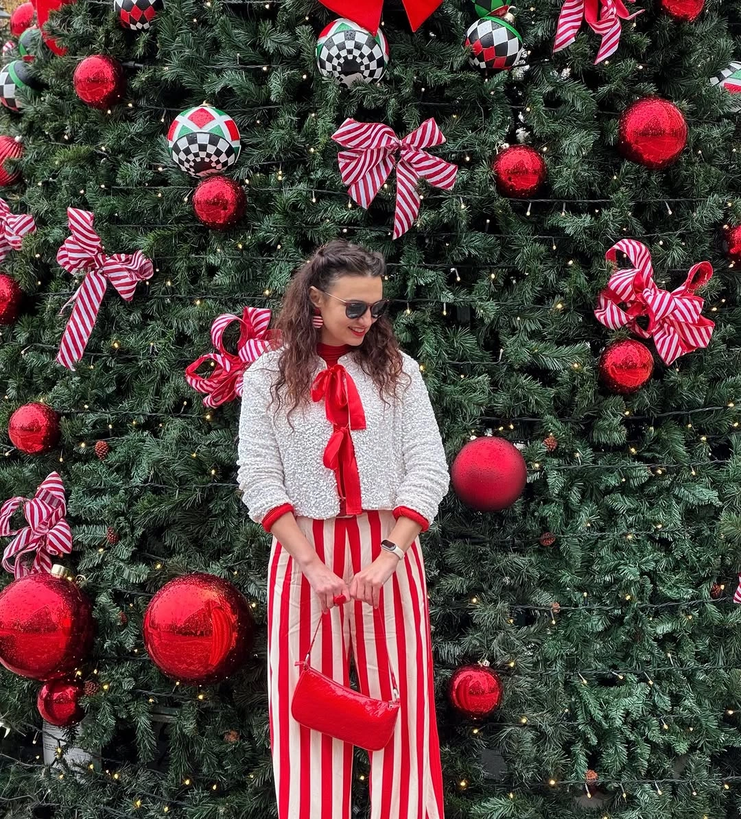 Red-and-white striped trousers, textured cardigan, bow blouse, and red handbag by Christmas tree