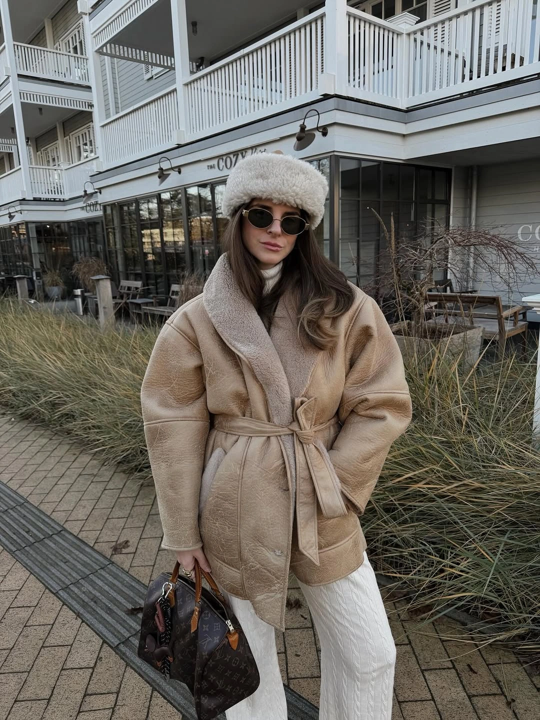 Belted taupe jacket, white trousers, pale hat, sunglasses, and dark handbag
