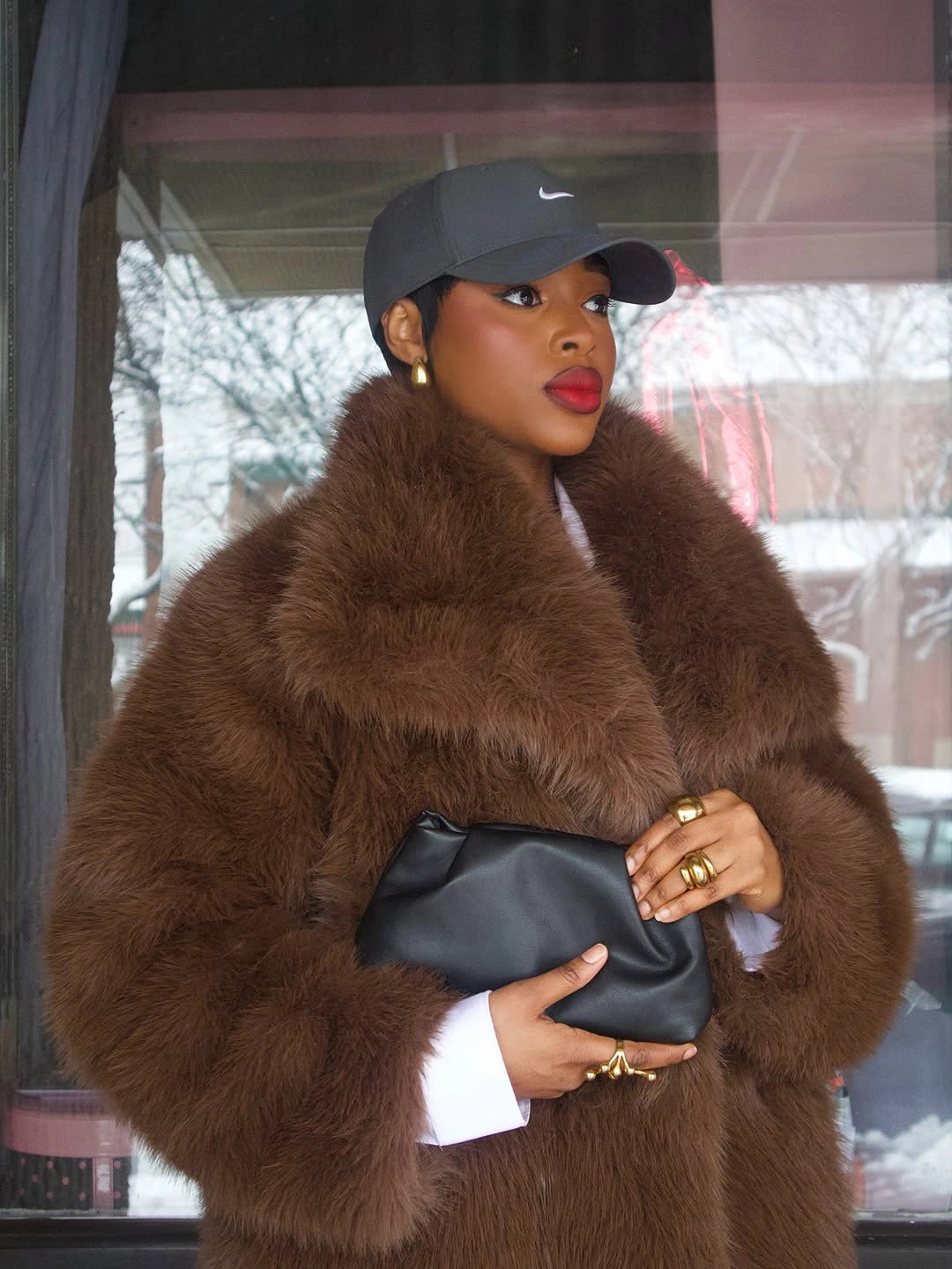 Brown shaggy coat, black cap, white cuffs, black clutch, jewelry, and red lips