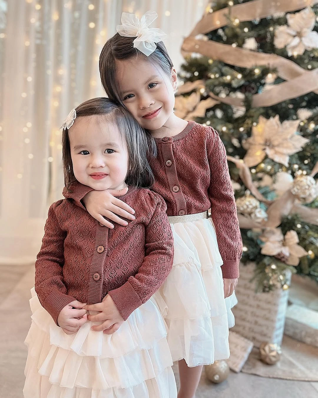 Two children in burgundy cardigans and layered white skirts