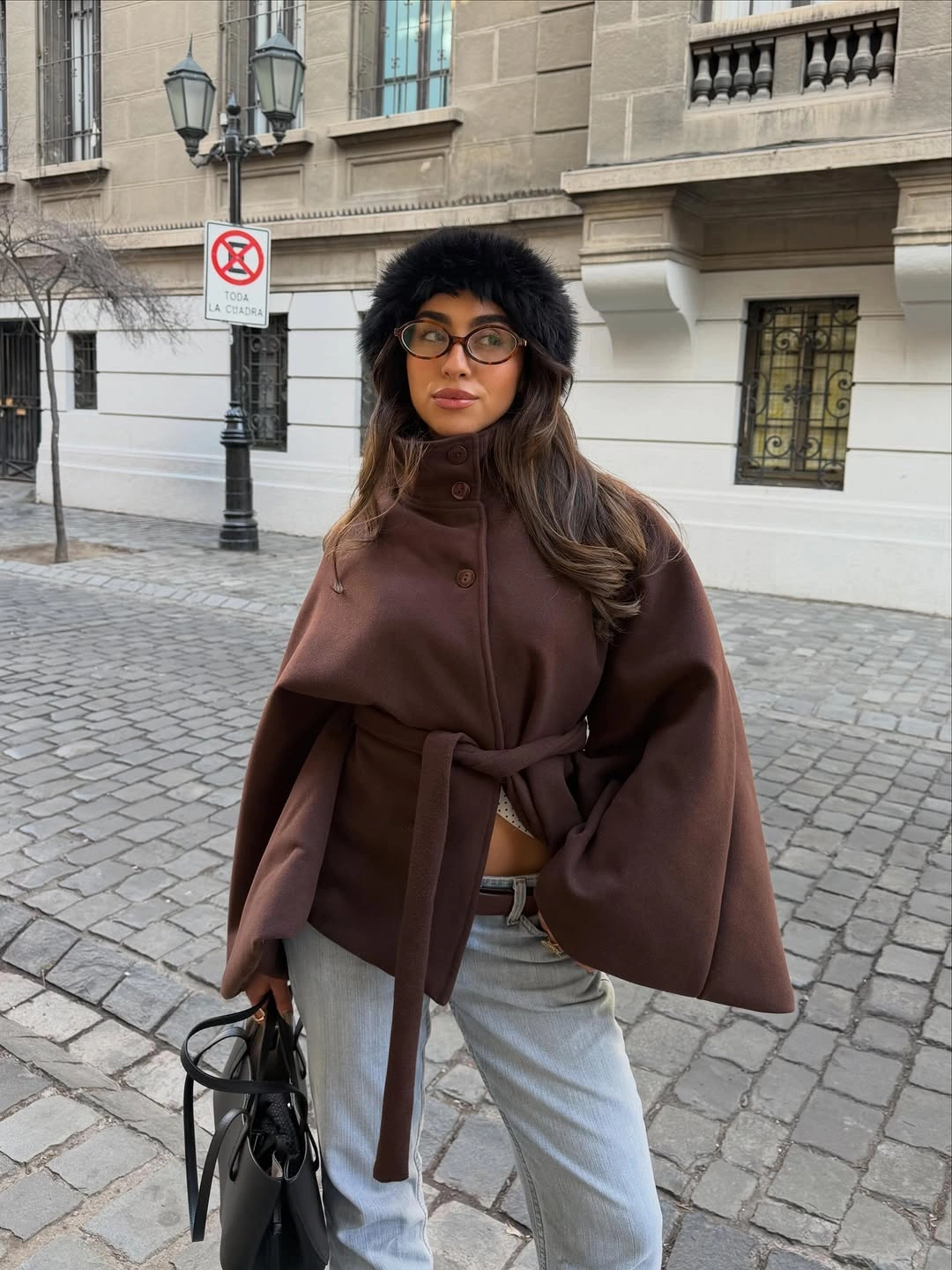Brown belted cape with pale jeans, black hat, and black handbag