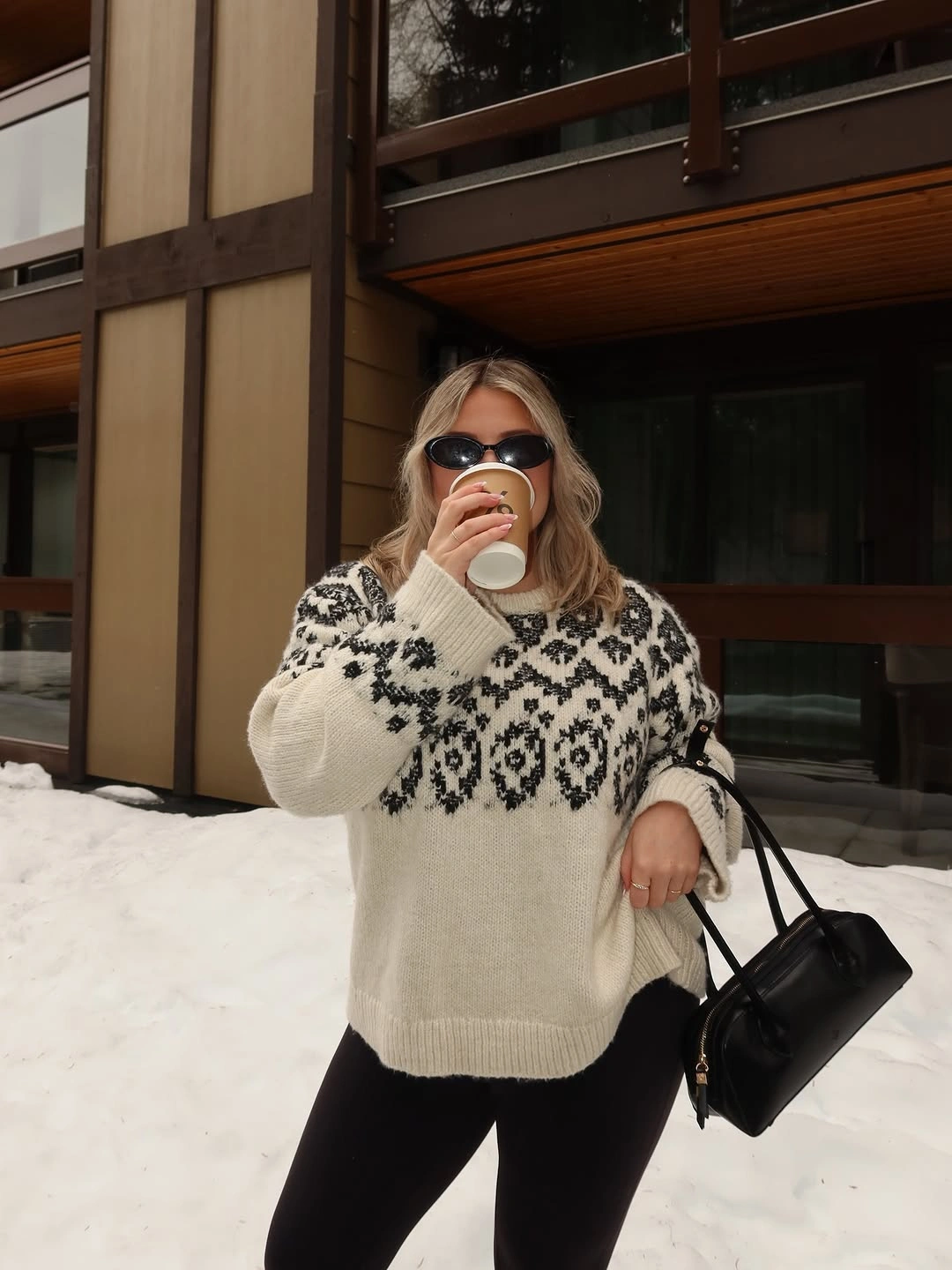 Cream patterned sweater with black pants, handbag, sunglasses, and snowy background