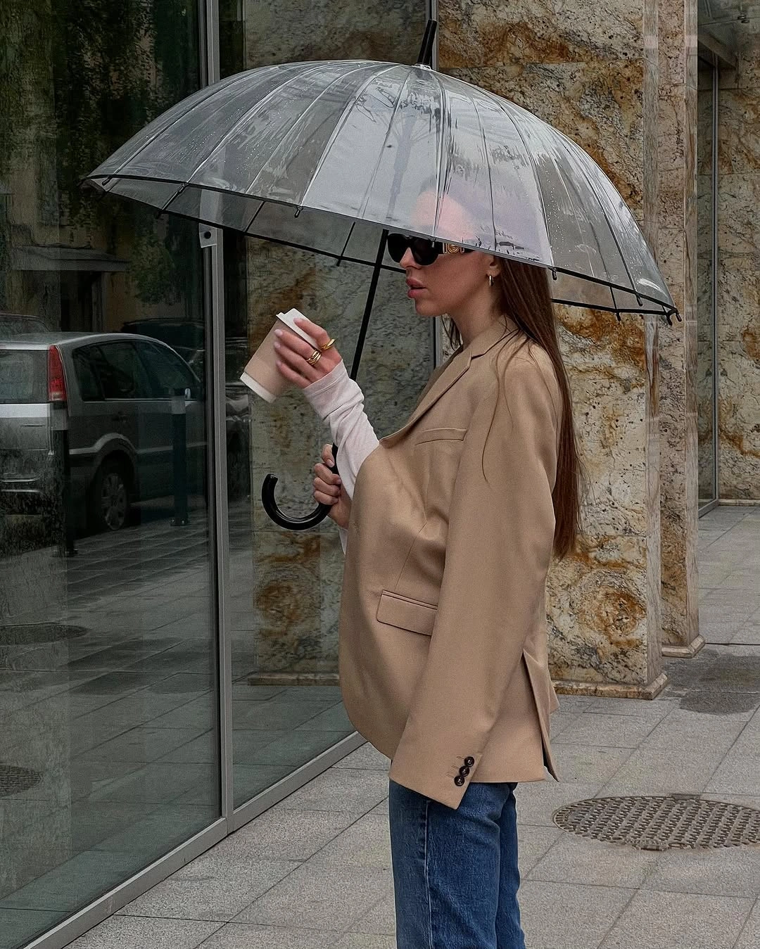 Woman in beige blazer, jeans, sunglasses, and clear umbrella