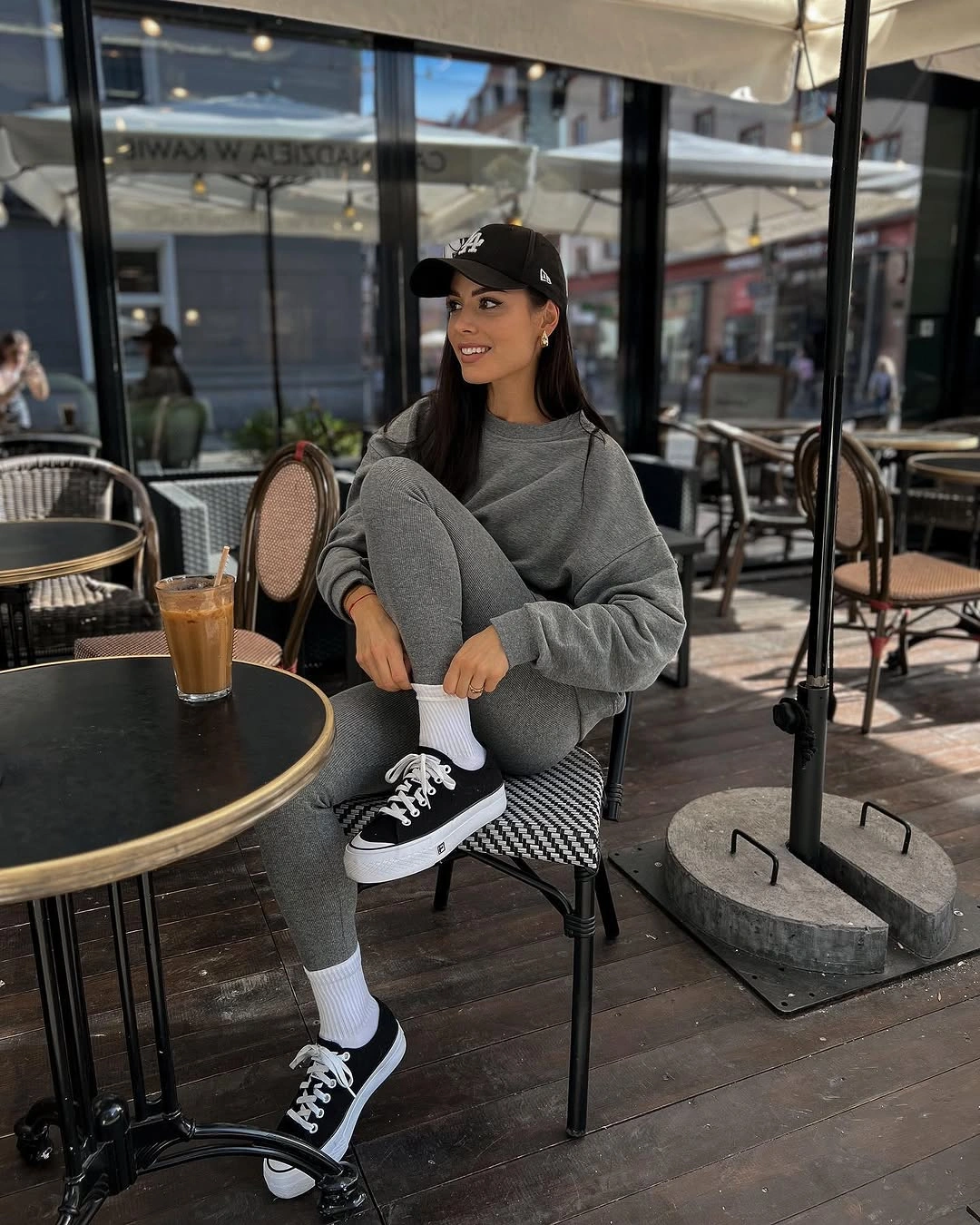 Seated woman in grey sweatshirt, joggers, black cap, and sneakers