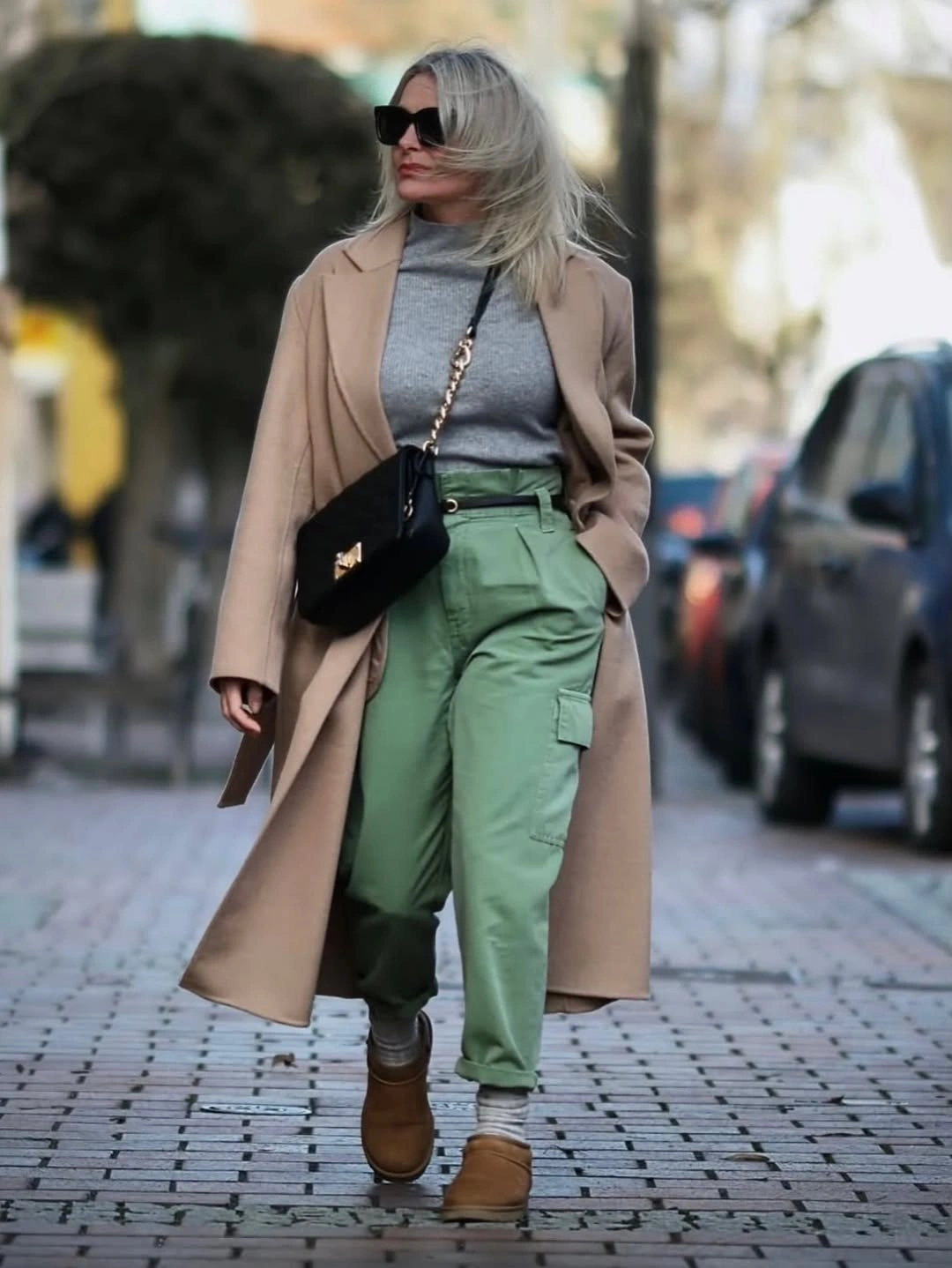Woman in camel coat, grey knit, green cargo trousers, and boots