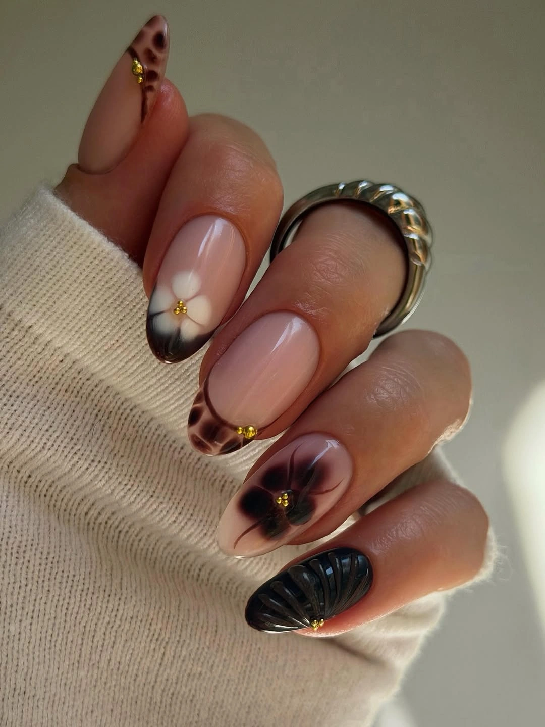 Long nude almond nails with brown patterns, gold beads, and black ridges