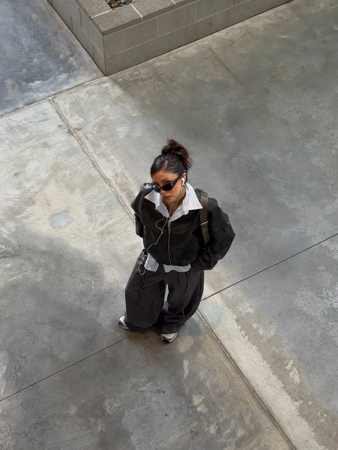 Dark jacket, white collar, loose dark trousers, sunglasses, and sneakers