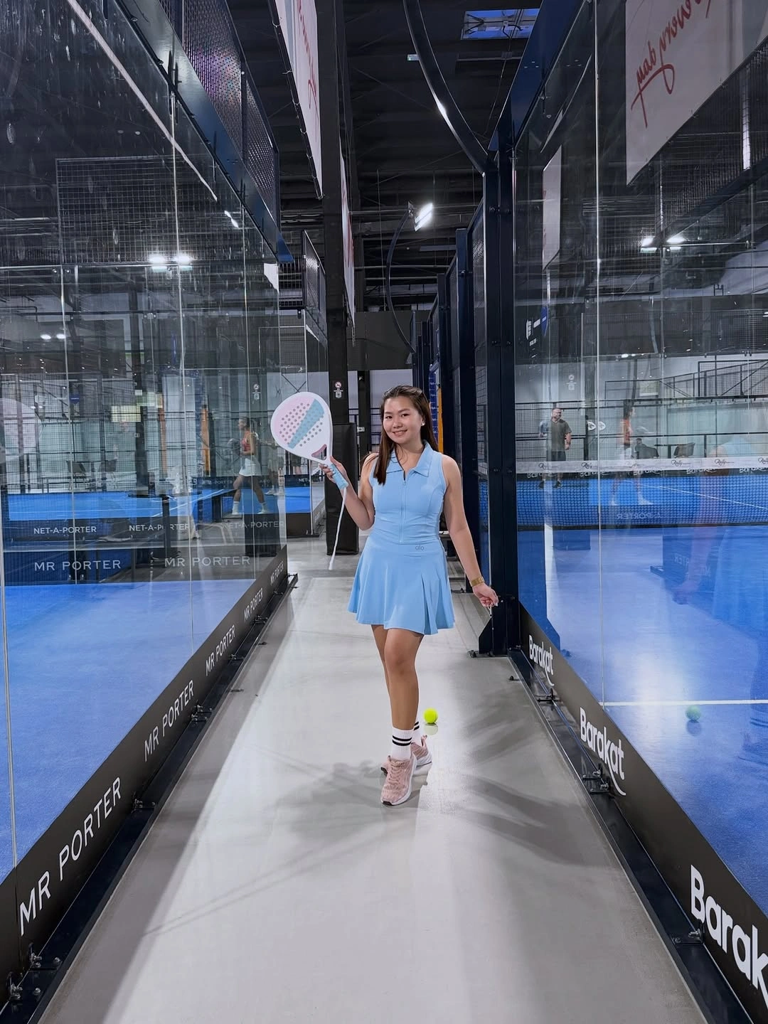 Light blue sleeveless padel dress carrying racket in glass court
