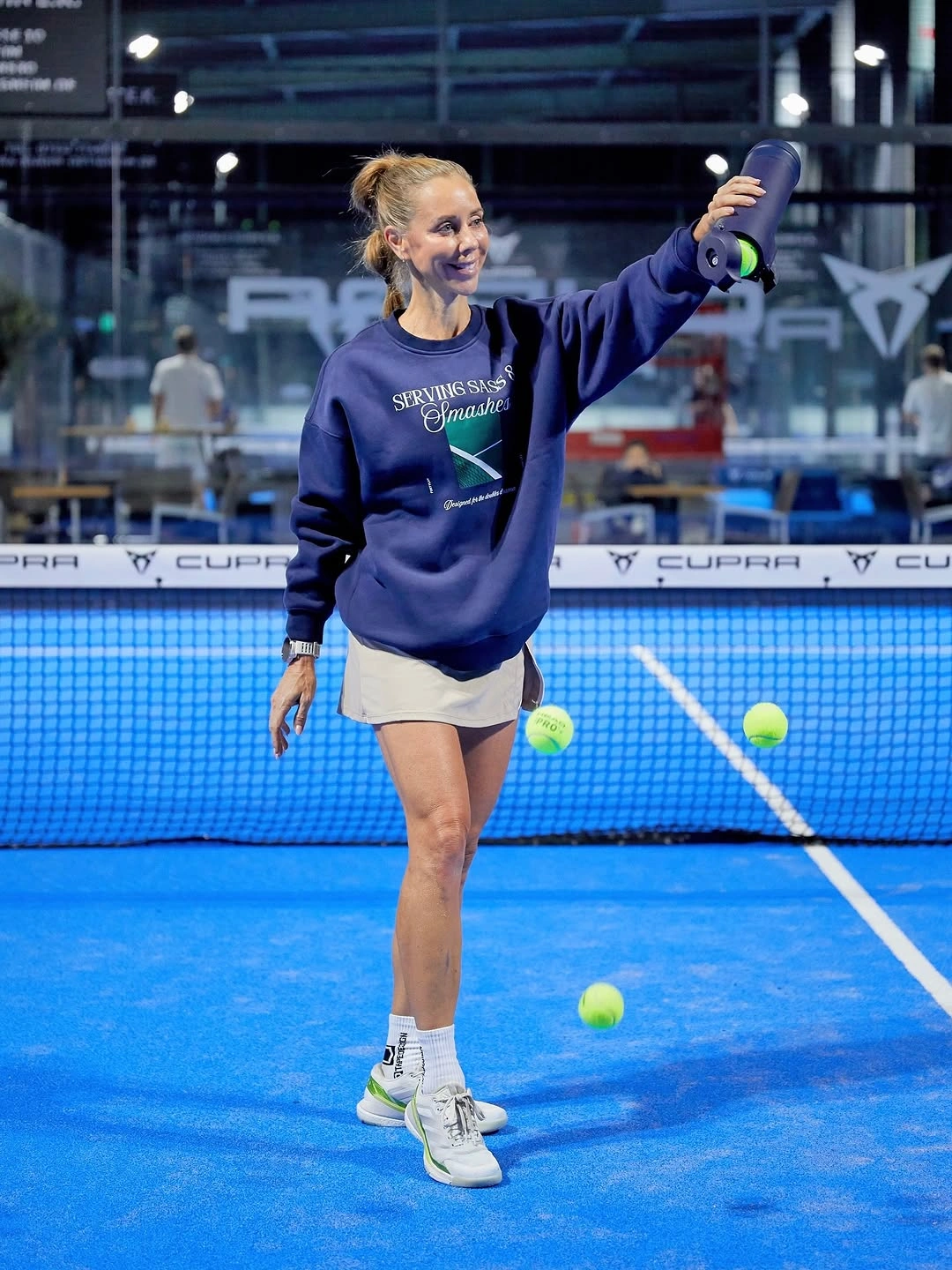 Cobalt sweatshirt over white skirt swinging padel racket indoors