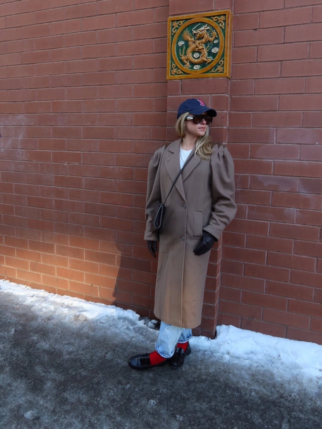 Long camel coat with cuffed pale jeans, dark cap, and red loafers