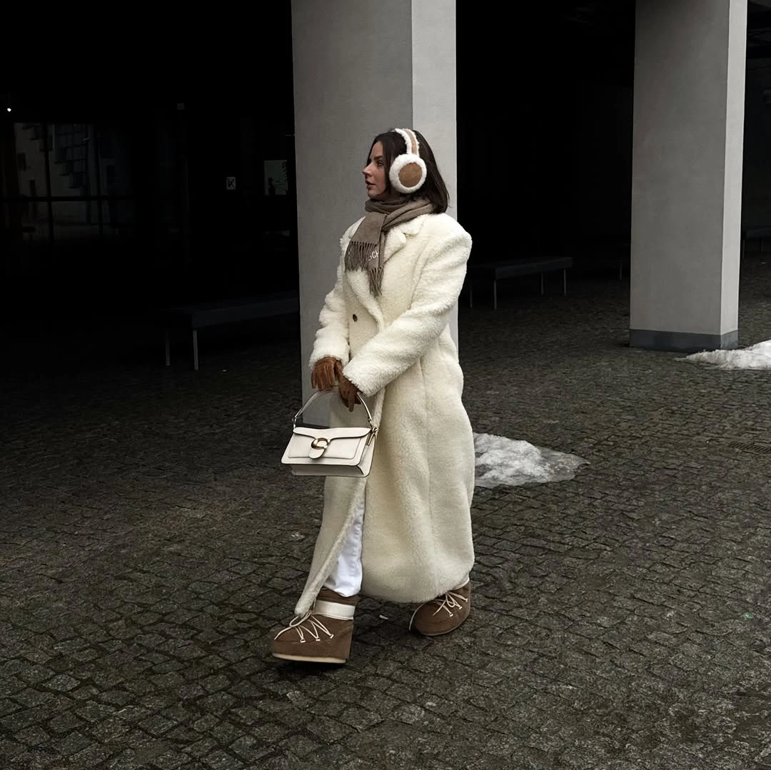 Long cream textured coat over patterned brown trousers and earmuffs