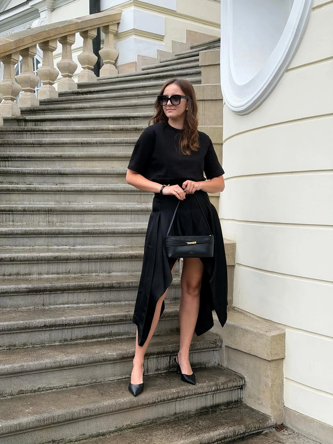 Black short-sleeve top, asymmetric draped skirt, pointed heels, and small handbag