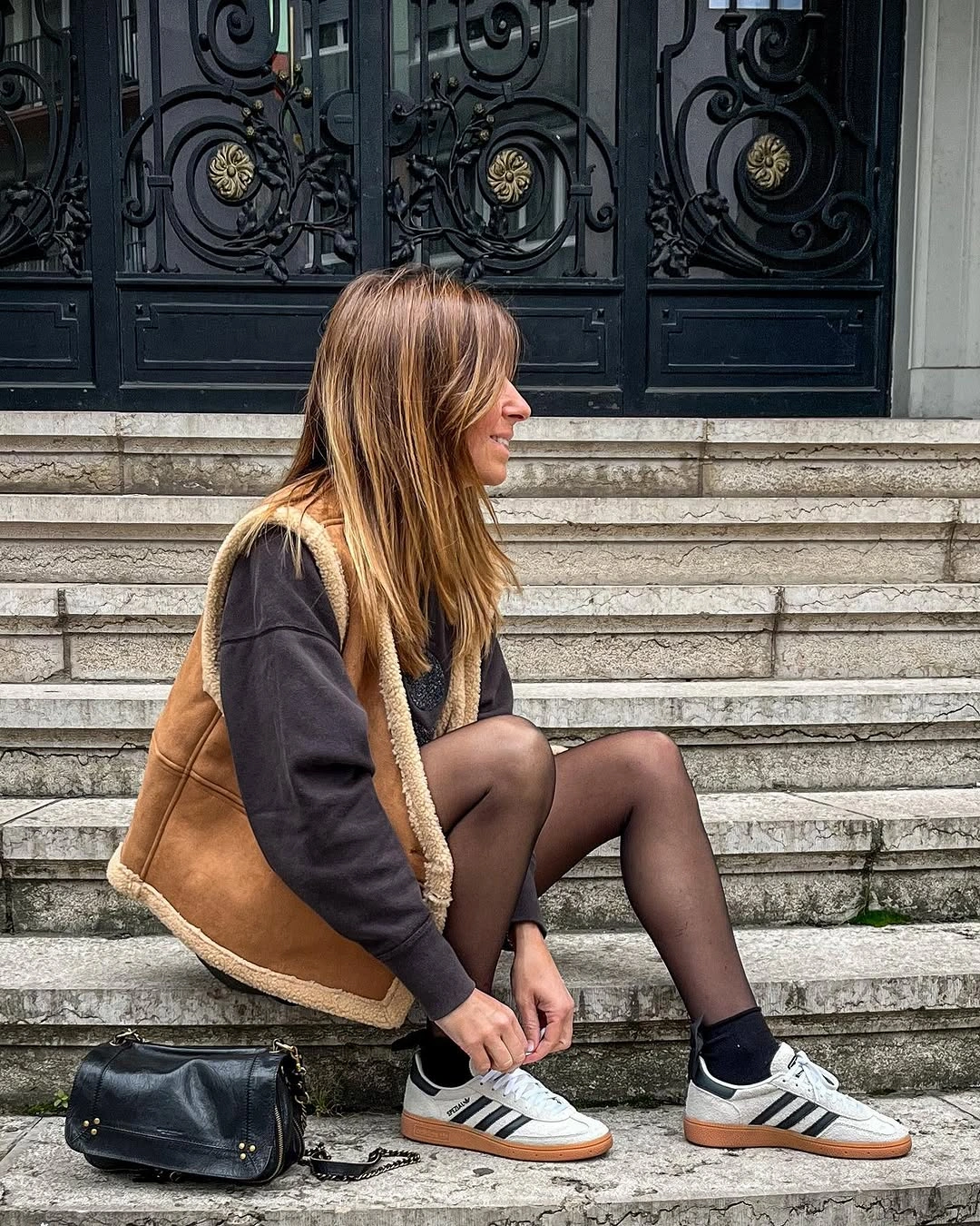 Tan trimmed vest, dark top, sheer tights, and white striped sneakers