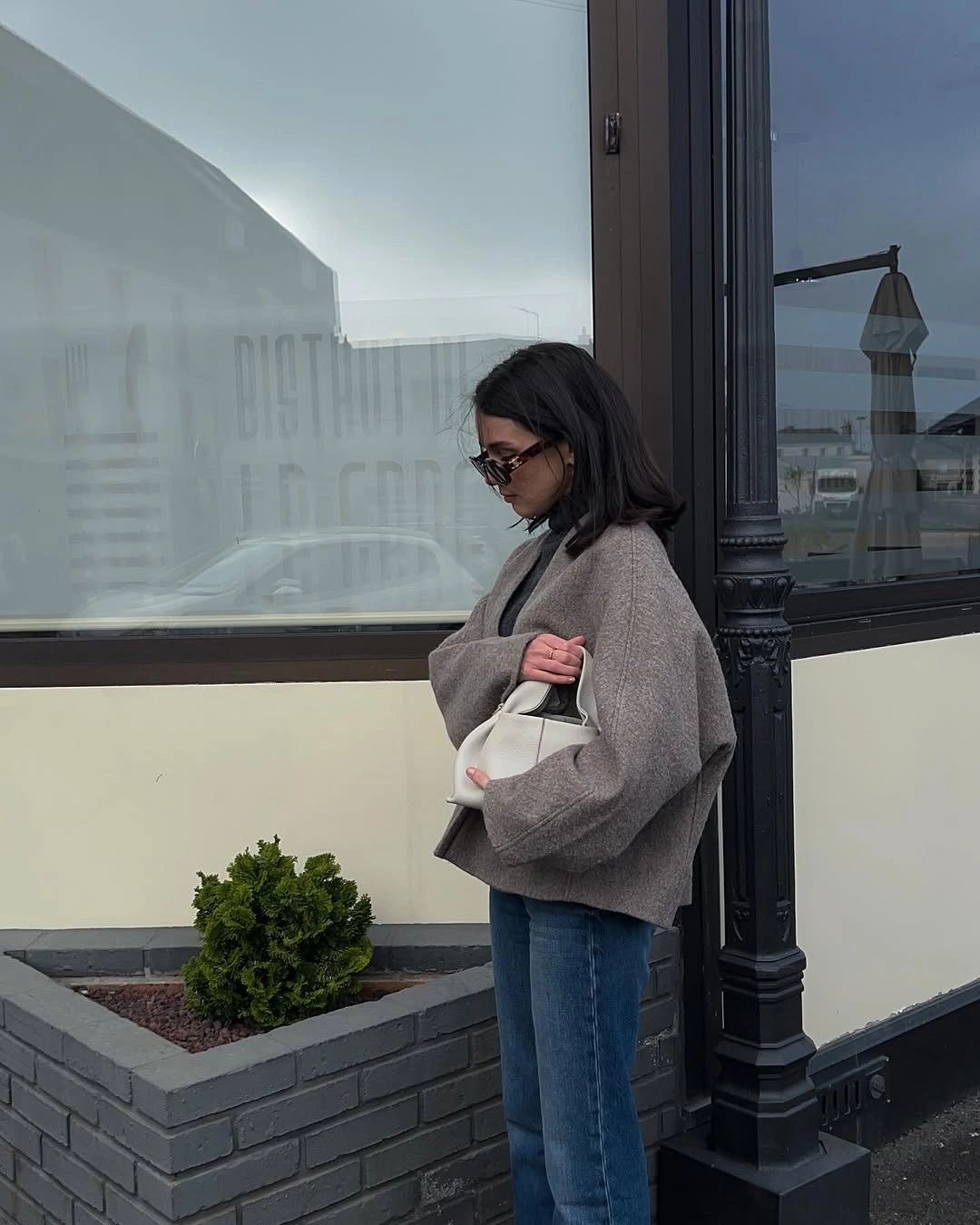 Oversized taupe-gray top with blue jeans, pale handbag, and sunglasses