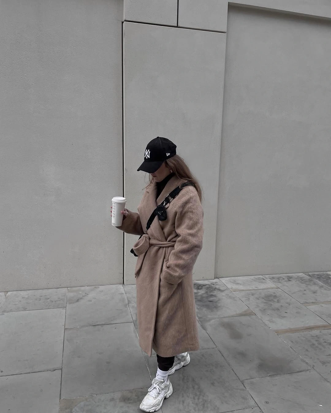 Long taupe belted coat with black cap, dark socks, and pale sneakers