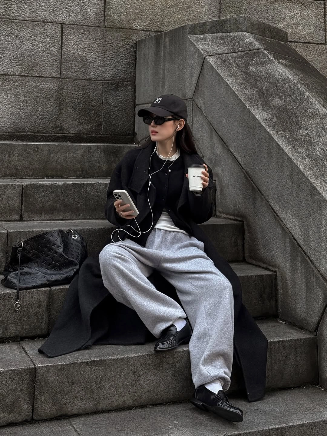 Black outer layer with gray sweatpants, dark loafers, cap, and quilted bag