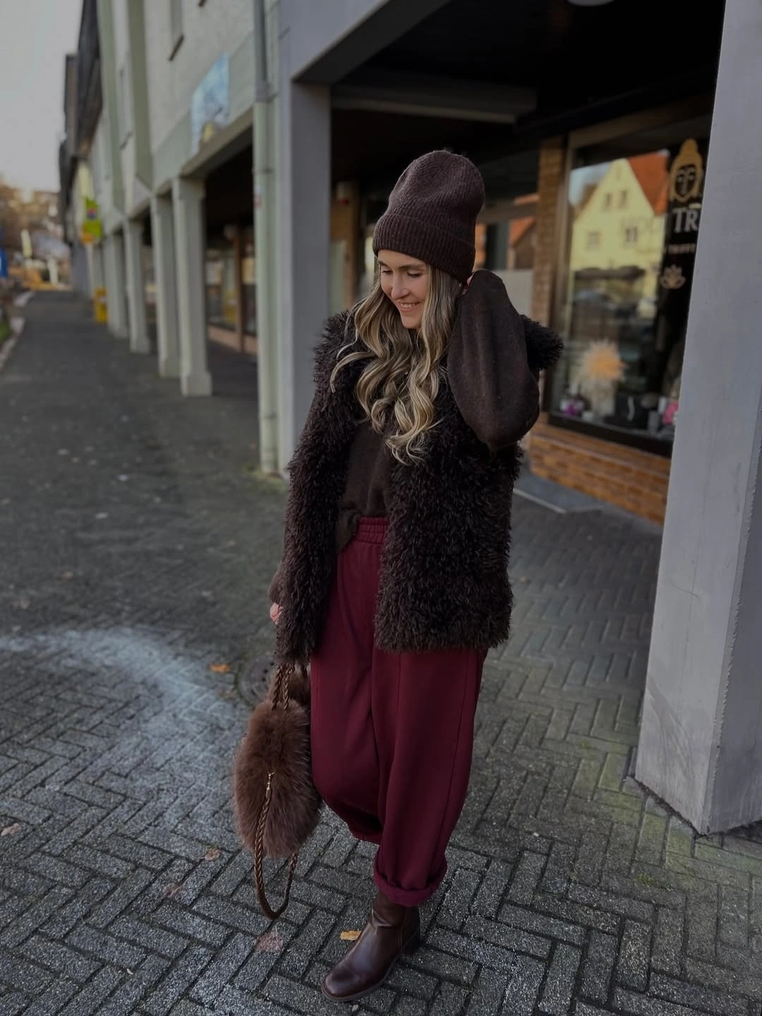 Dark shaggy vest, burgundy trousers, brown boots and knit beanie