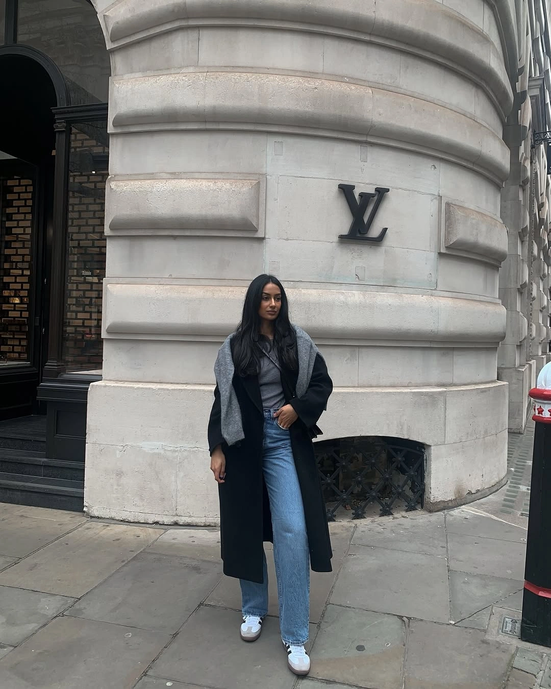 Long black coat with gray shoulder layer, blue jeans, and white sneakers