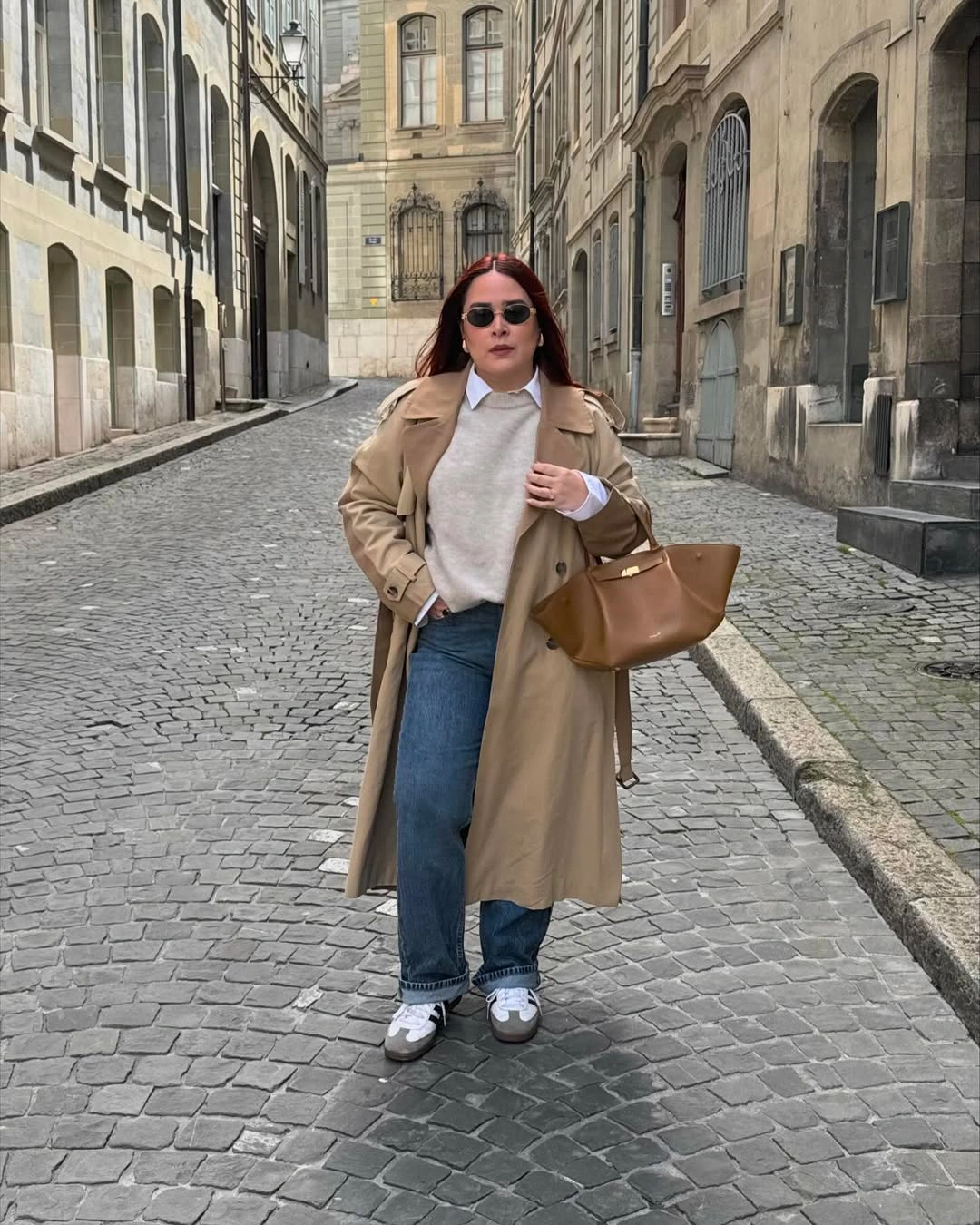 Beige trench coat over gray sweater with blue jeans and brown handbag