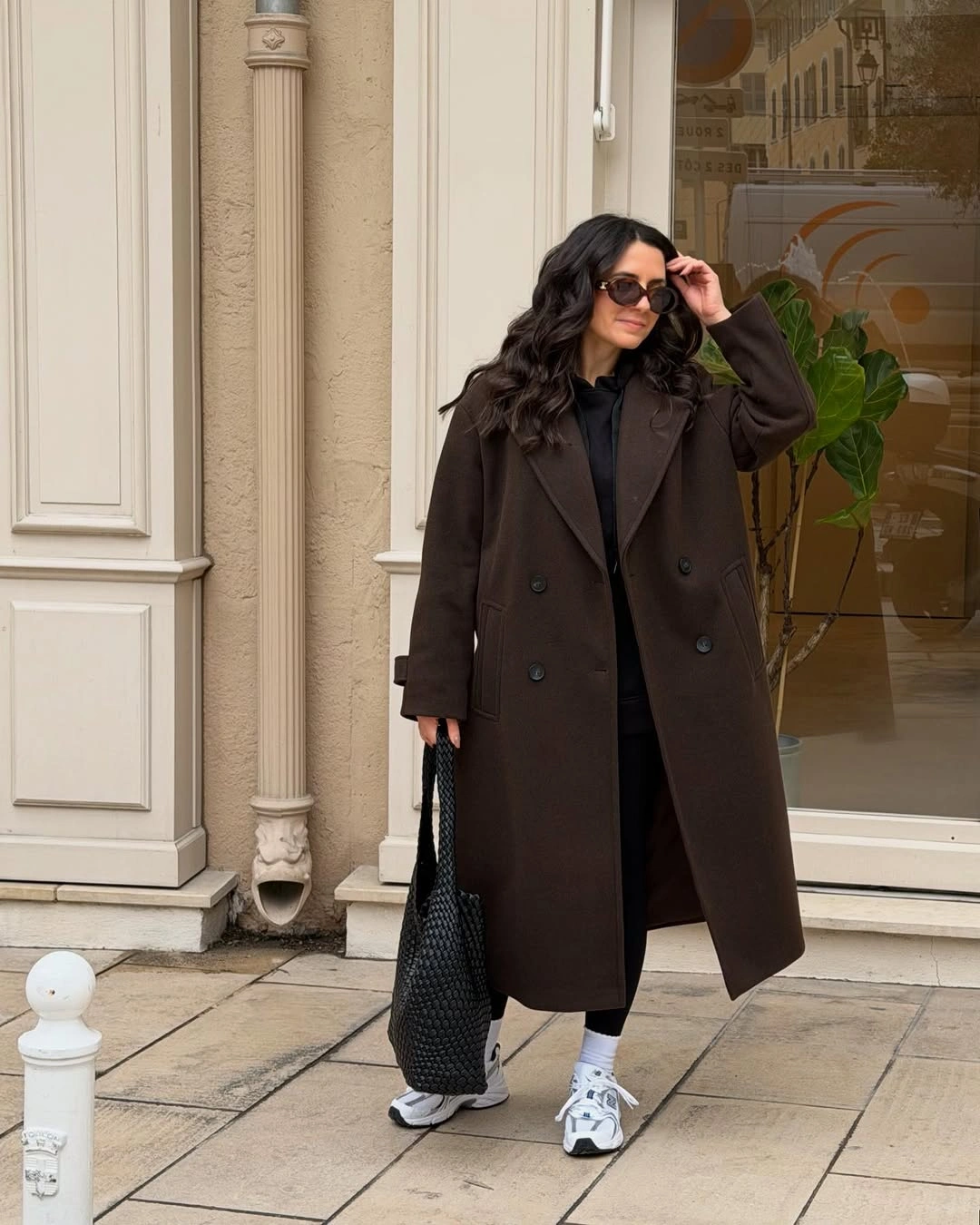 Long dark brown coat with black tote, white socks, and gray sneakers