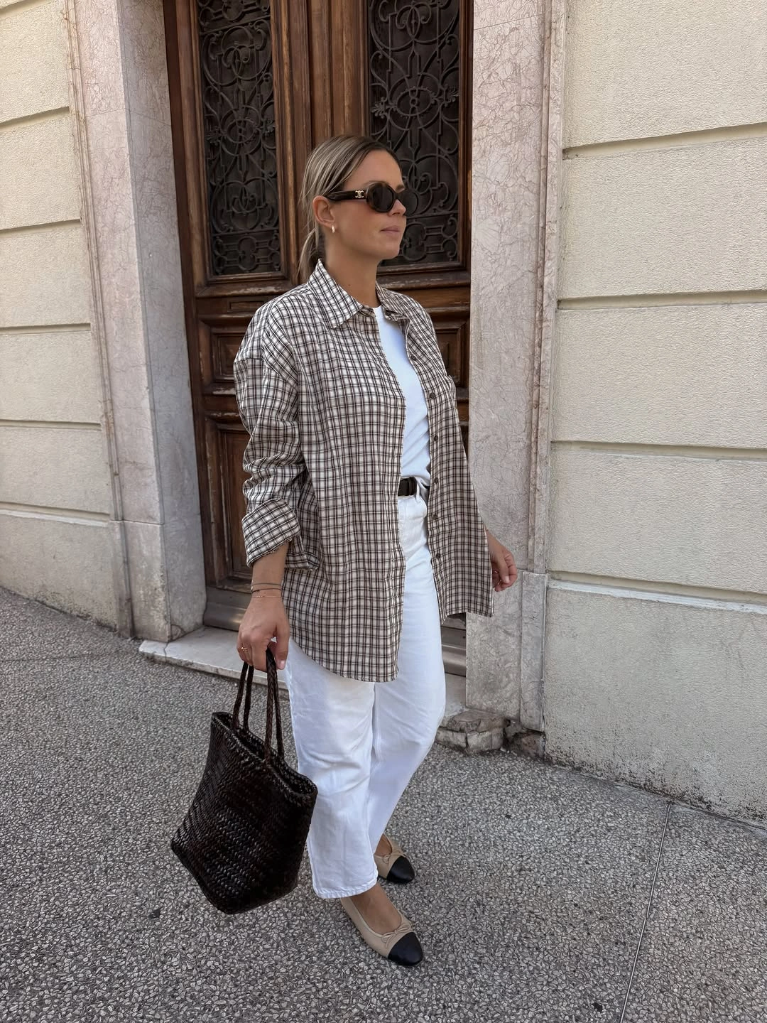 Brown checked overshirt with white trousers, woven dark tote, and two-tone flats