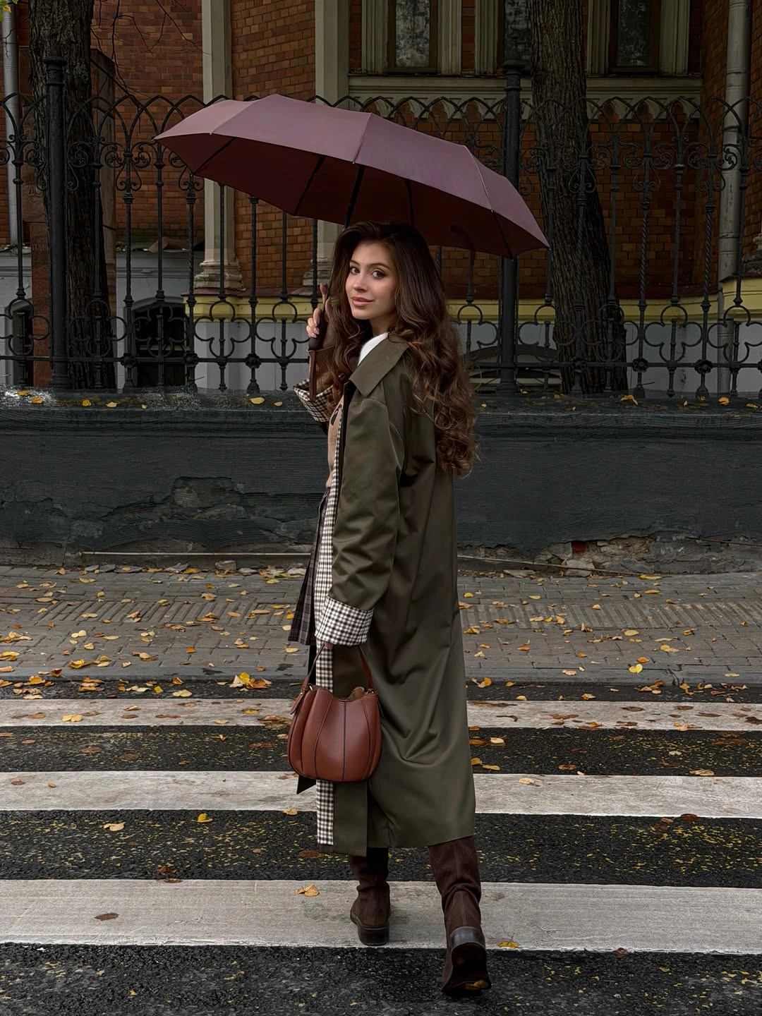 Long olive coat with checked details, brown boots, and burgundy umbrella