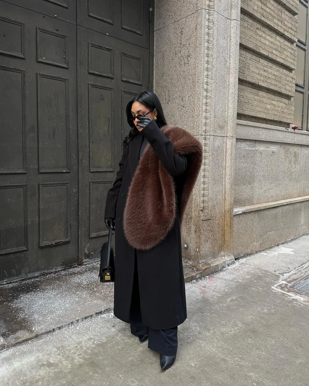 Long black coat with brown shaggy shoulder drape, dark trousers, and pointed footwear