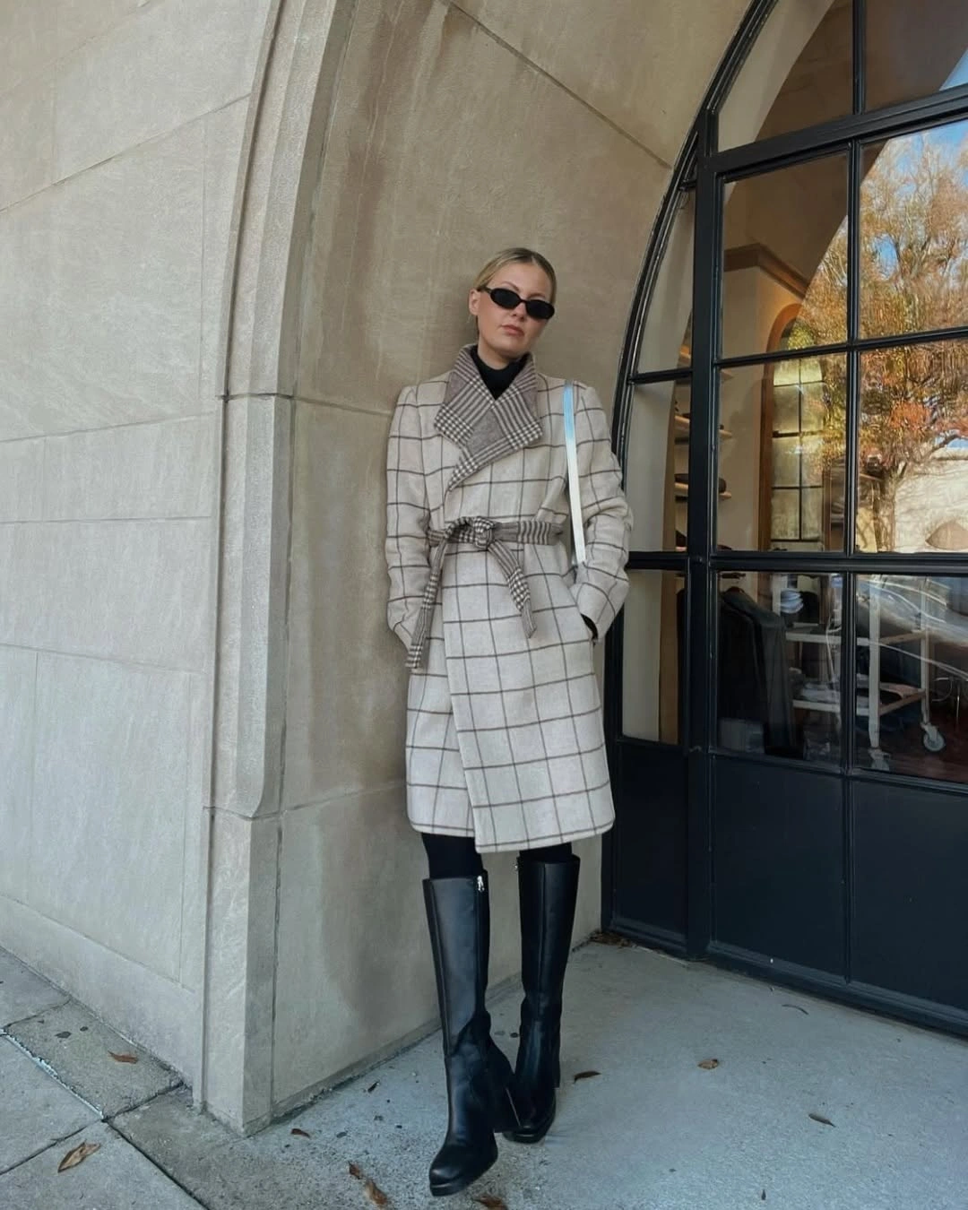 Long ivory checked coat with tied belt, black top, and tall boots