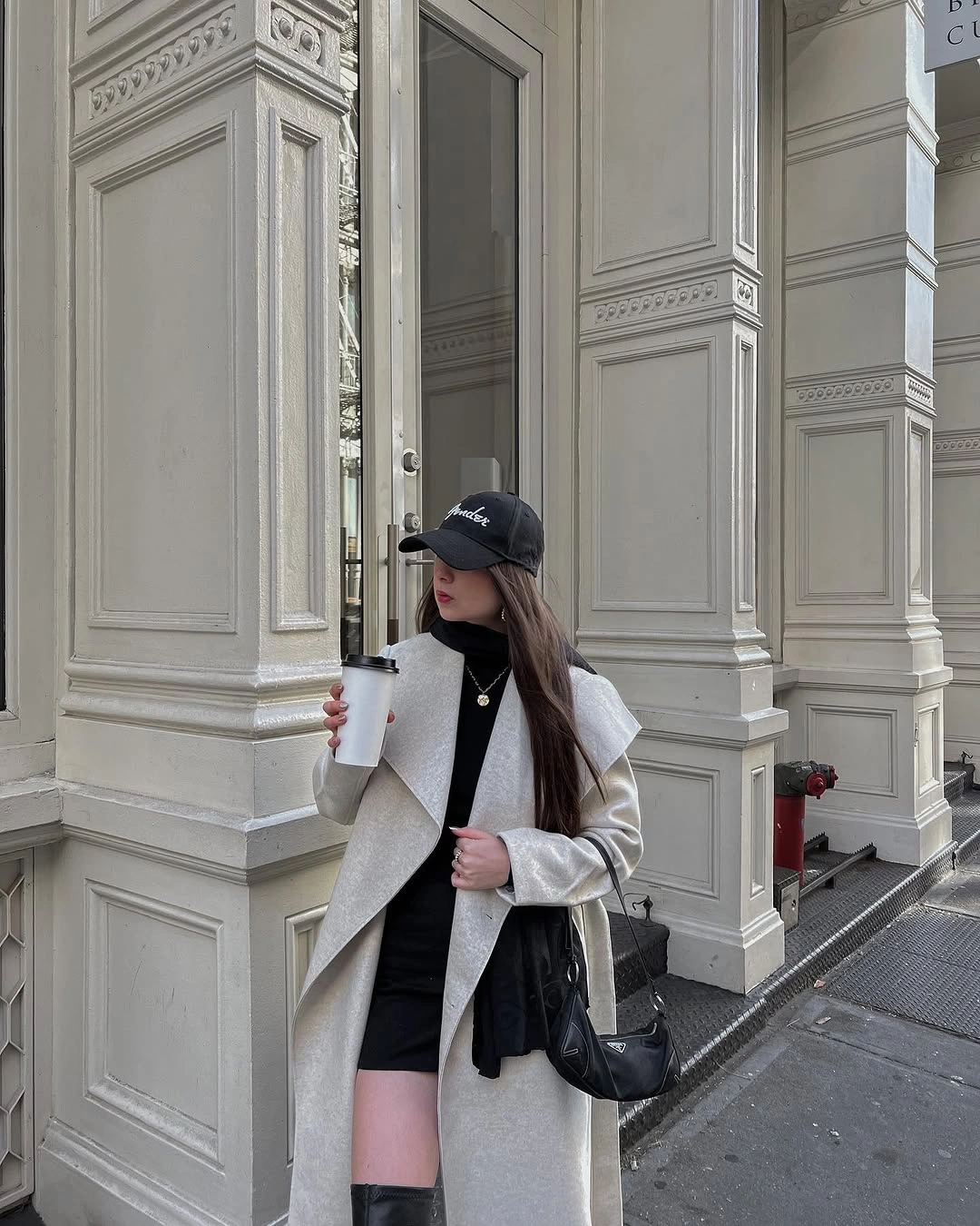 Long ivory coat, black high-neck layer, tall boots, and baseball cap