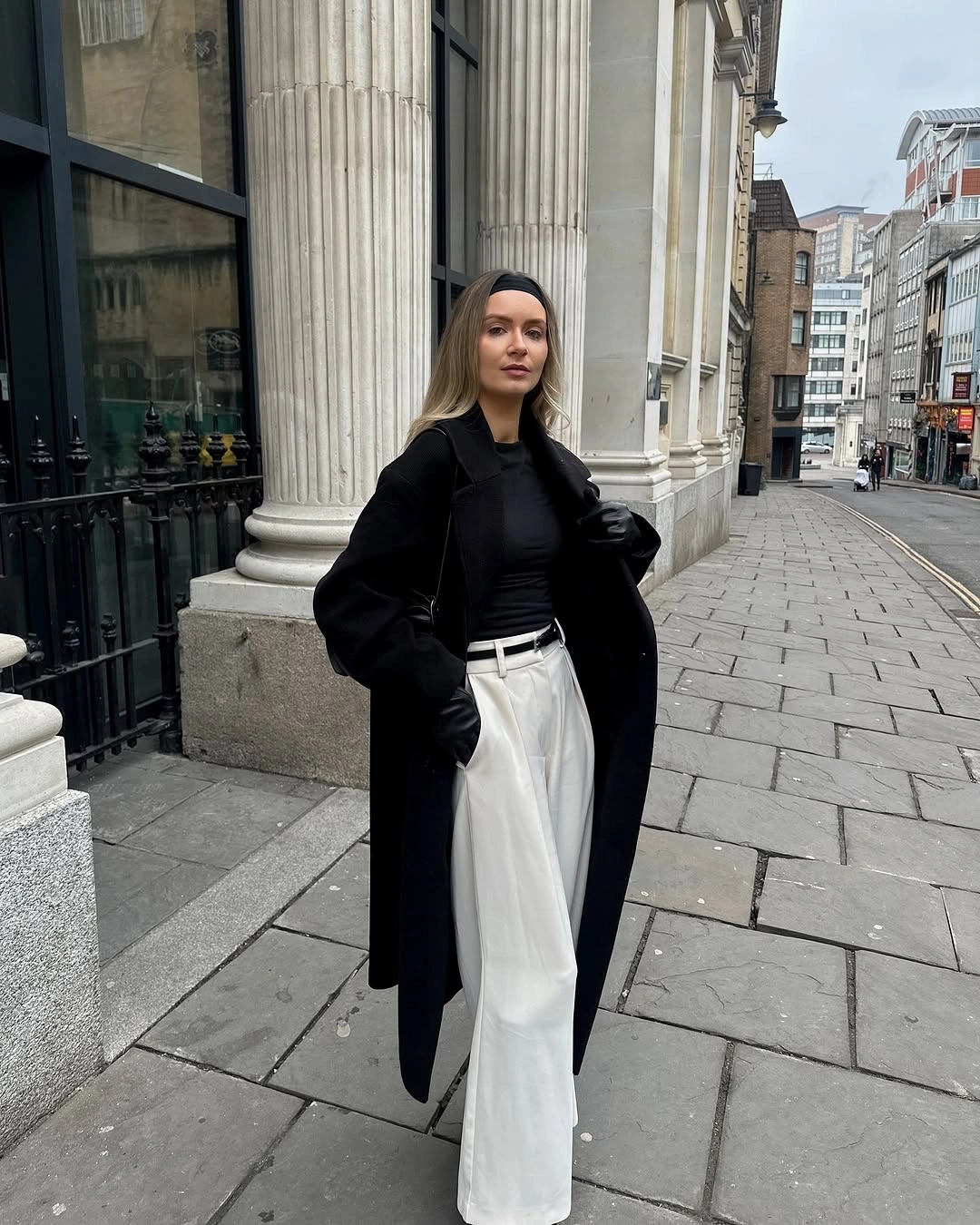 Long black coat, fitted black top, white pleated trousers, and black gloves
