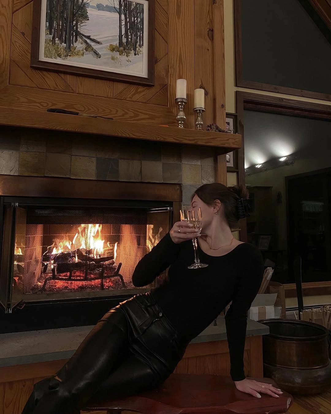 Fitted black long-sleeved top and glossy dark trousers beside fireplace