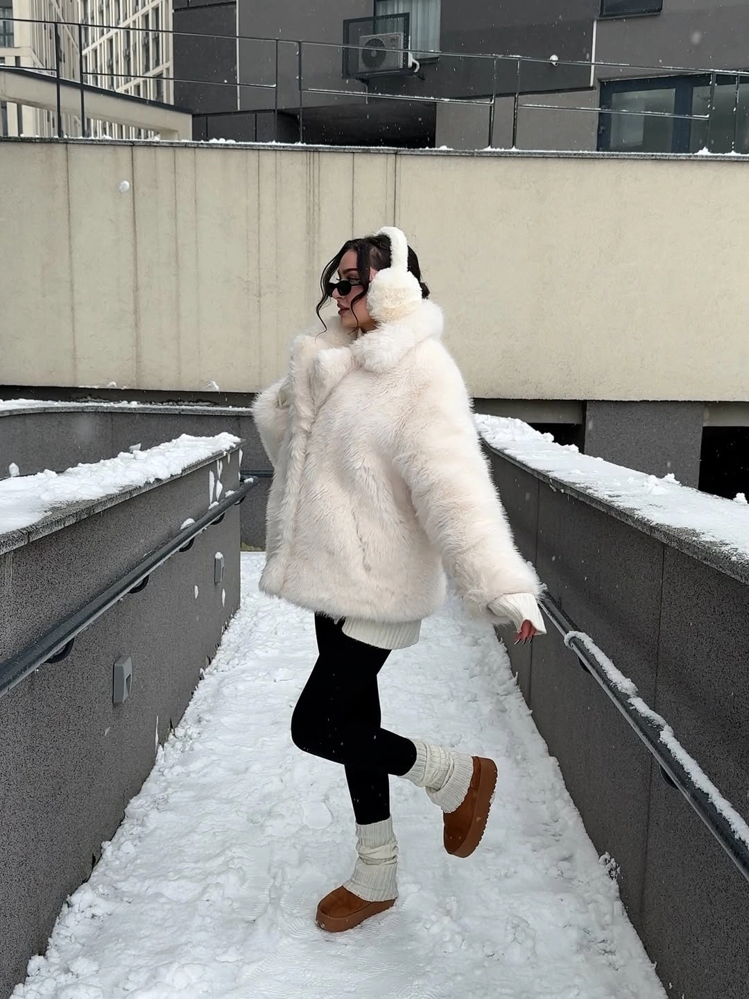 Pale fluffy jacket, earmuffs, black leggings, and tan boots in snow