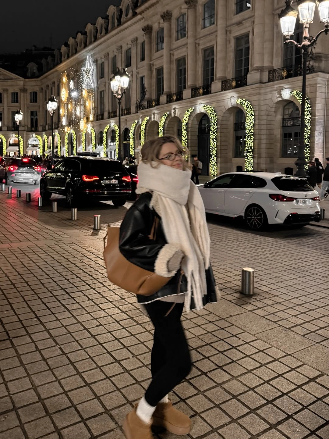 Short black jacket with oversized ivory scarf, fitted black bottoms, and tan boots