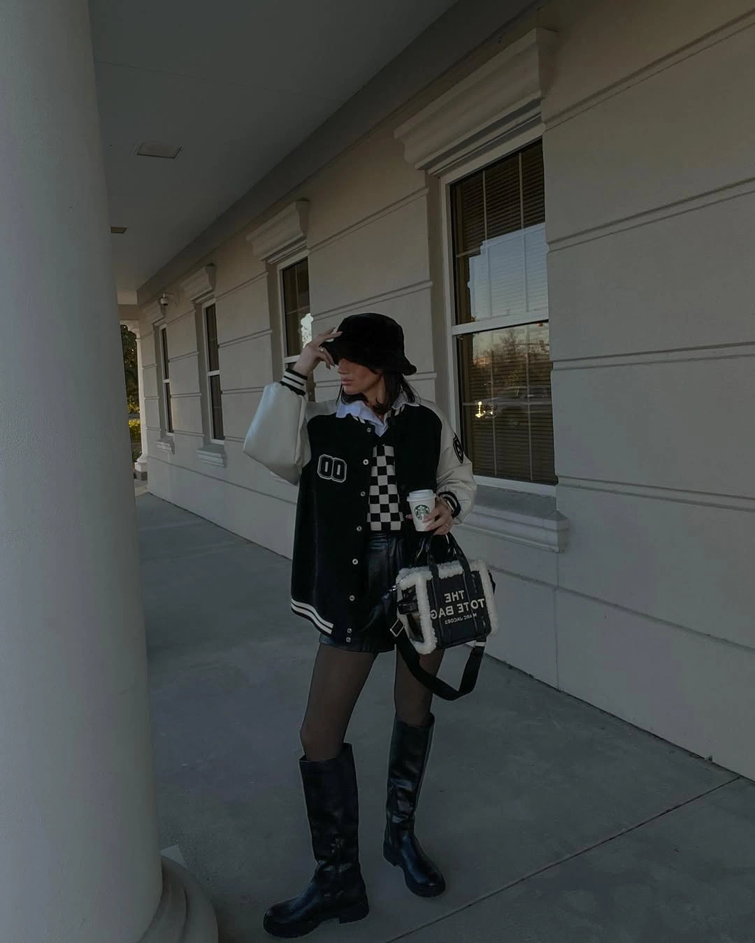 Woman wearing black varsity jacket, dark bucket hat, tights, and tall boots.