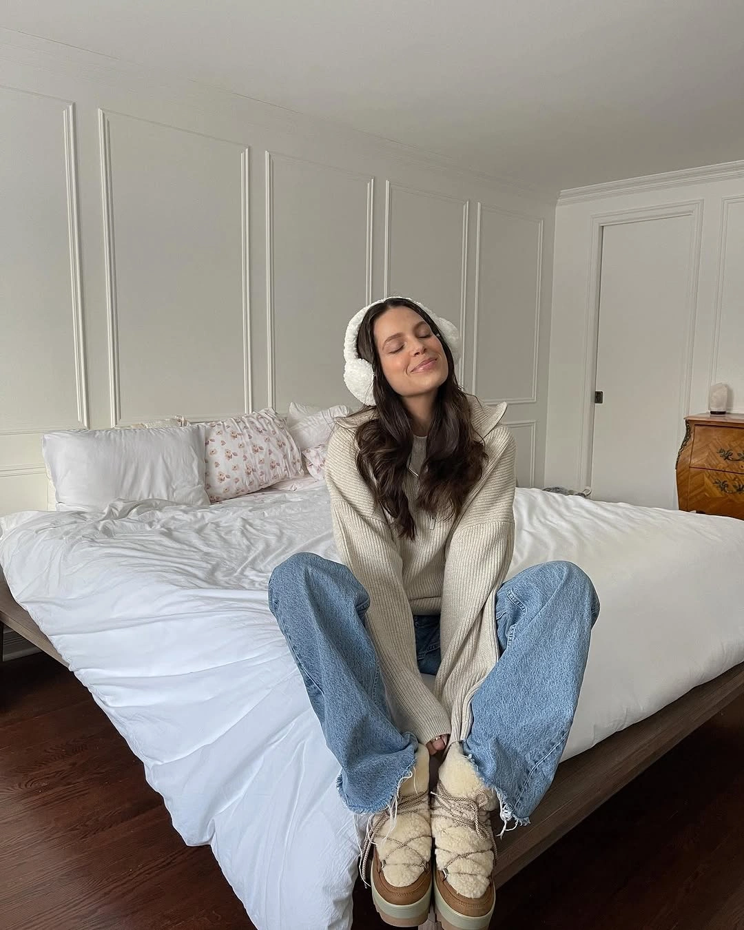Oversized pale knit top, wide light blue jeans, earmuffs, and lace-up shoes.