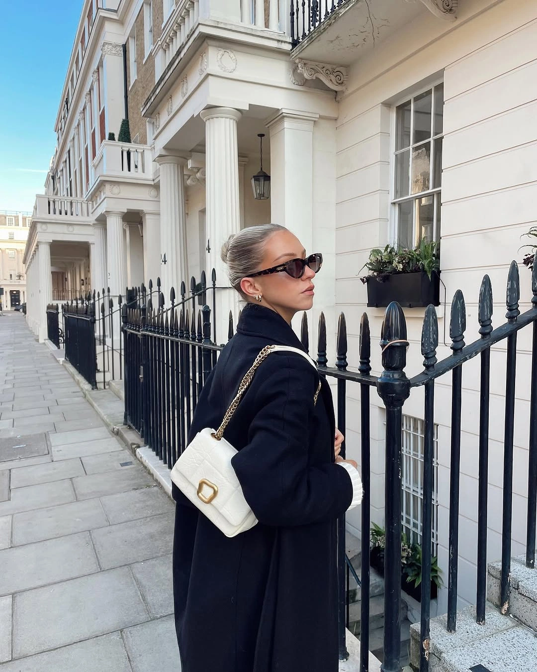 Dark tailored coat, ivory chain-strap handbag, sunglasses, and pulled-back hair.